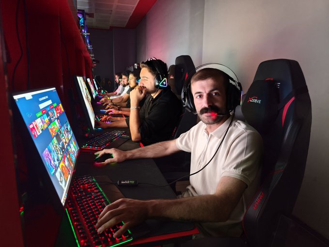 A man wearing a headset and beige polo shirt sits at a gaming PC in a LAN center, with a glowing RGB keyboard and monitor displaying game thumbnails, while other gamers are visible in the background.