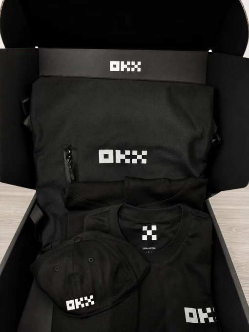 An open OKX merchandise box containing branded apparel including a black duffel bag, t-shirt, and cap, all featuring the OKX logo in white.