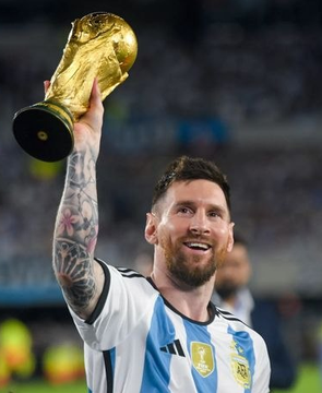 Lionel Messi, wearing Argentina's light blue and white striped jersey, joyfully hoists the FIFA World Cup trophy after winning the 2022 World Cup in Qatar.