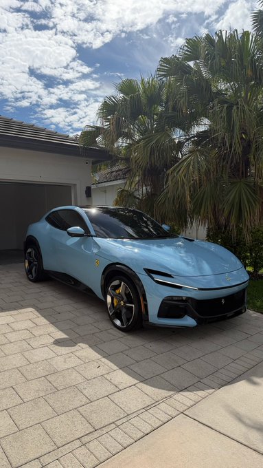 A light blue Ferrari Purosangue parked on a paved driveway in front of a house with palm trees.