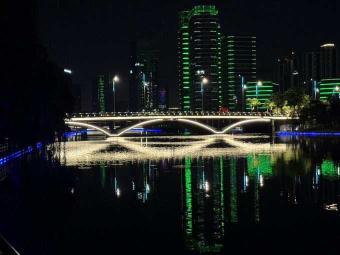 A nighttime cityscape shows a modern bridge with arched lights spanning a body of water, reflecting green and white lights from the surrounding skyscrapers.