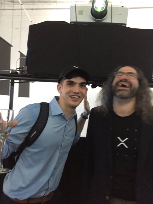 Two men pose together at what appears to be an event; the younger man on the left wears a black baseball cap with a white X logo and a light blue shirt, while the older man on the right has long gray hair, a beard, and wears a black jacket with the same X logo.