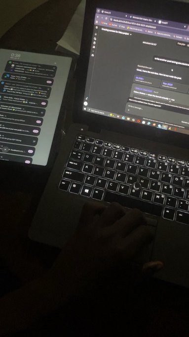 A dark photo of a hand on a laptop trackpad next to a tablet displaying a messaging app, with a wedding ring visible on the person's finger.