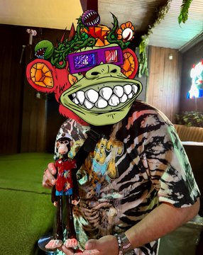 A person wearing a vibrant tie-dye shirt holds a uniquely designed, colorful figurine or sculpture. The artwork features a large green grinning face wearing sunglasses that display the words 'TEKK' and 'PULP', topped with a miniature top hat and surrounded by tropical and whimsical elements.