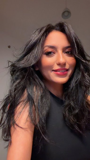 A selfie portrait of a young woman with long, wavy dark hair wearing a black top, smiling slightly at the camera.