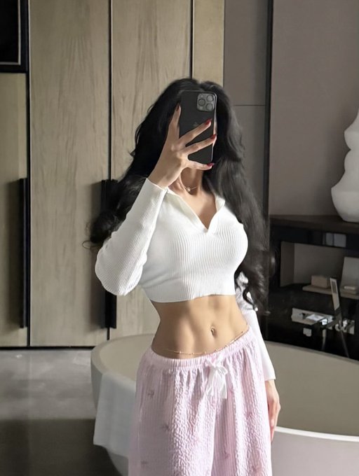 A woman takes a mirror selfie in a modern room, wearing a fitted white long-sleeve crop top that reveals her midriff, paired with pink textured shorts. The stylish outfit and relaxed morning vibe match the casual "Gm" greeting.