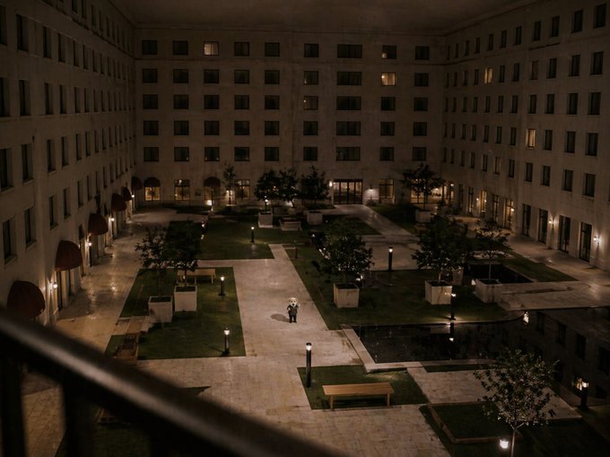 The image shows an empty courtyard with several potted plants, trees, and benches scattered throughout. A person can be seen walking through the courtyard at night. The scene appears to be a part of a movie poster for "Water Faucet Pattern".