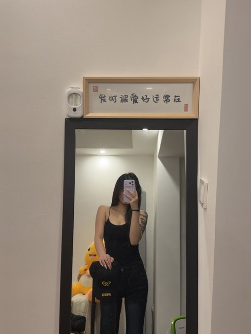 A mirror selfie of a woman in a black tank top and dark jeans, with a Chinese calligraphy sign above the mirror and a plush duck and MCM bag visible in the background.