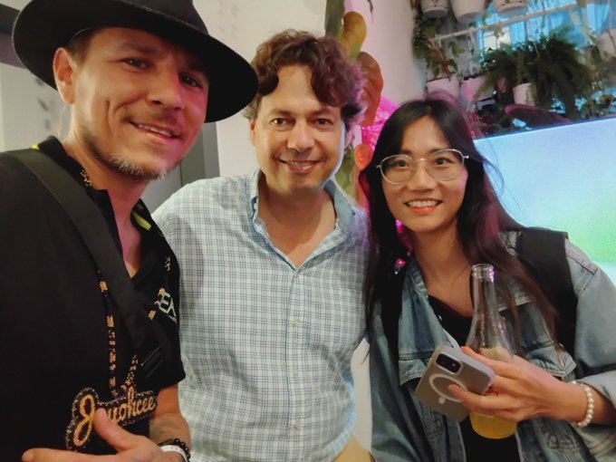 Three smiling people pose together for a photo at what appears to be a social event or party, with the man on the left wearing a black hat and shirt reading 'Jiu-tstice'.