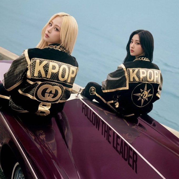Two K-pop style women wearing matching black and gold jackets with 'KPOP' lettering, sitting on a maroon vintage car with 'FOLLOW THE LEADER' written on the side, by a body of water.