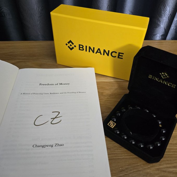 A signed copy of "Freedom of Money" by Changpeng Zhao (CZ), founder of Binance, displayed alongside a yellow Binance-branded gift box and a black bracelet in a Binance-branded case, commemorating the Binance Campus Asia 2026 event.