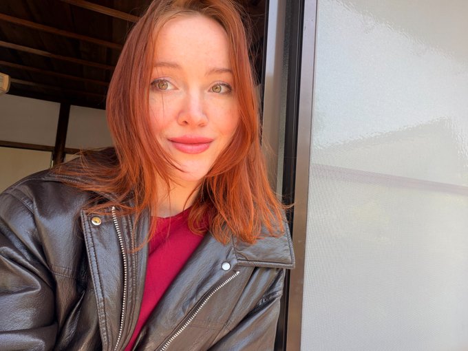 A young woman with long red hair, wearing a black leather jacket over a red top, takes a selfie in front of a window.