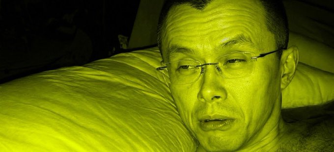 A close-up selfie of a man wearing rimless glasses lying on a pillow, with the entire image tinted in a strong yellow-green color.