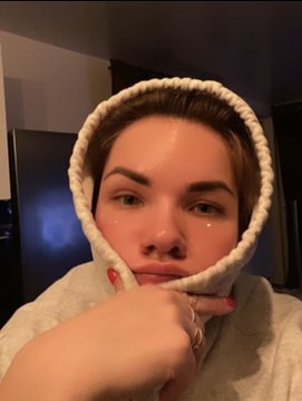 A young woman wearing a cream-colored knitted hood takes a close-up selfie, resting her chin on her hand adorned with a ring and a tear-shaped sticker. The cozy, intimate portrait complements the "GM" (Good Morning) post, likely serving as a personal morning greeting to accompany the NFT listing link for "Sweet danger" on objkt.com.