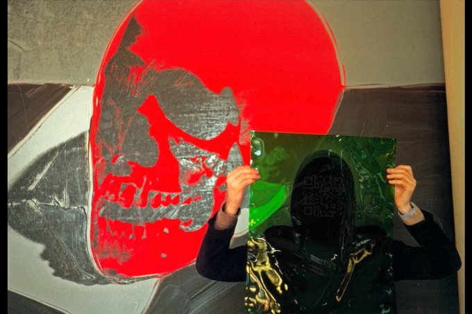 An image of a person holding a green transparent sheet with a QR code, standing in front of a red and black stencil portrait resembling Andy Warhol's style.