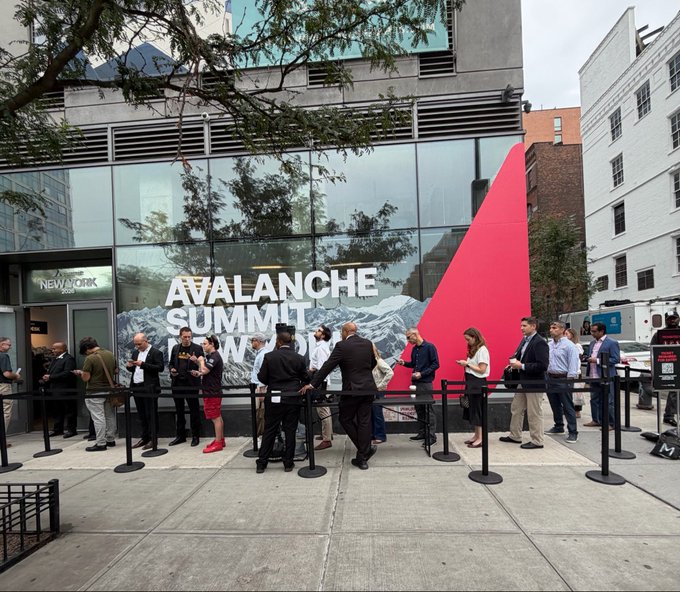 Attendees line up outside the New York venue where large window signage advertises the "AVALANCHE SUMMIT NEW YORK," a blockchain industry event being covered live on social media.