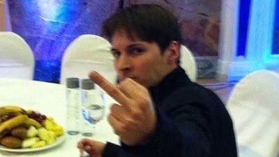 A man in a dark shirt sits at a table with a plate of food and water bottles, raising his middle finger toward the camera.