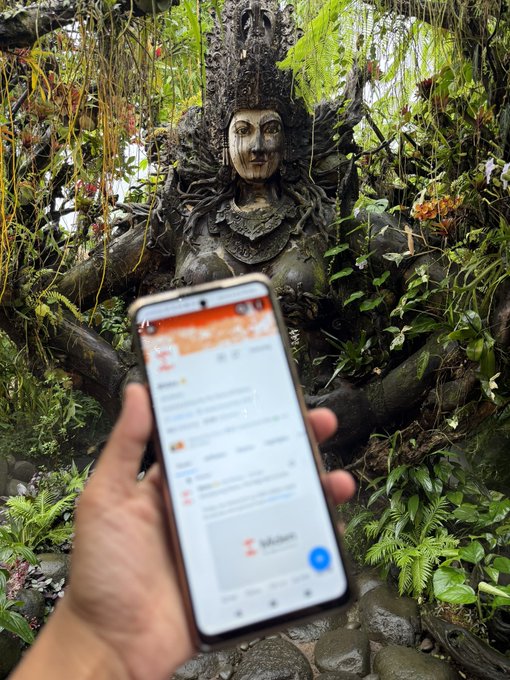 A hand holds a smartphone displaying a messaging app with text in Indonesian, set against the backdrop of a stone statue entwined with tree roots and surrounded by greenery.
