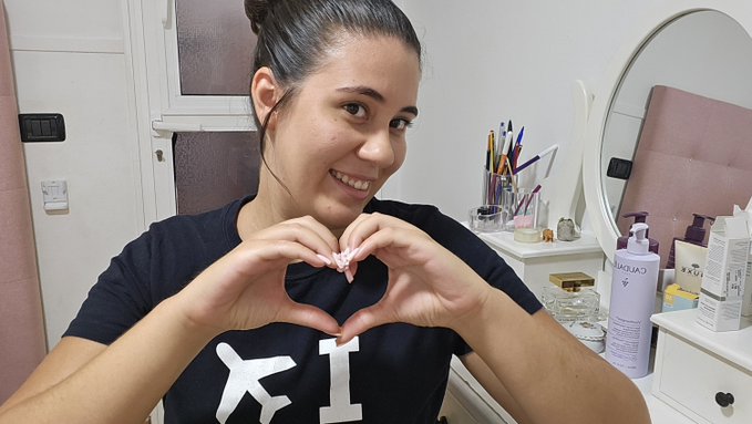 A woman in a dark blue shirt smiles at the camera while making a heart shape with her hands, sitting at a vanity table with a round mirror and various makeup products visible in the background.