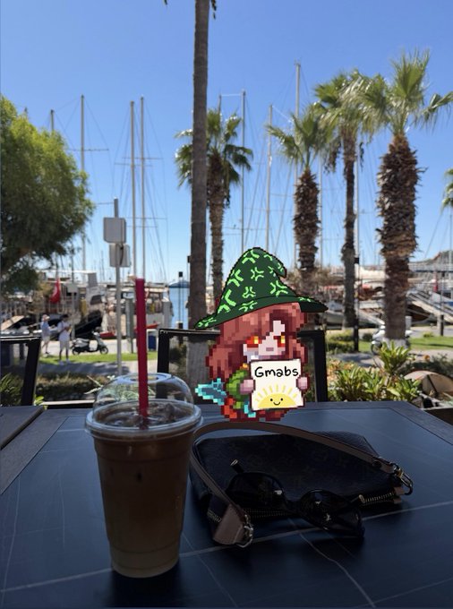 A photo of an iced coffee in a clear cup with a red straw sitting on a table next to sunglasses, with a pixel art overlay of a character wearing a green hat and holding a sign that says 'Gmabs', set against a background of palm trees and sailboats in a marina.