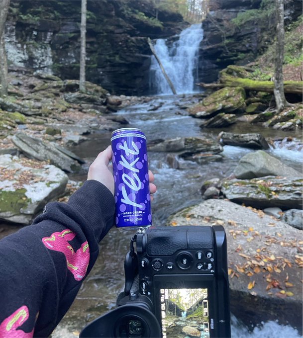 A hand holds up a purple can of "rekt" Moon Cookies beverage in front of a scenic waterfall backdrop, with a camera capturing the moment. The image showcases a collaboration between MoonPay and Rekt Drinks in a natural outdoor setting.