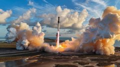 A SpaceX rocket launches from a pad, creating massive clouds of smoke and steam against a blue sky with scattered clouds.