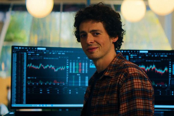 A young man with curly hair wearing a plaid shirt stands in front of two computer monitors displaying stock trading charts and data.