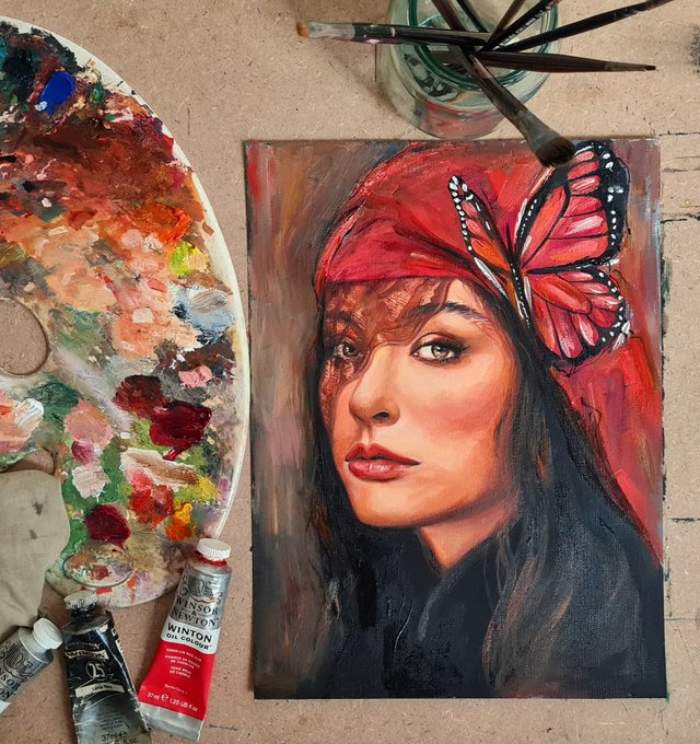 A painted portrait of a woman with dark hair wearing a red headwrap adorned with a large monarch butterfly, displayed alongside a paint palette, brushes, and a Winsor & Newton cadmium red paint tube.