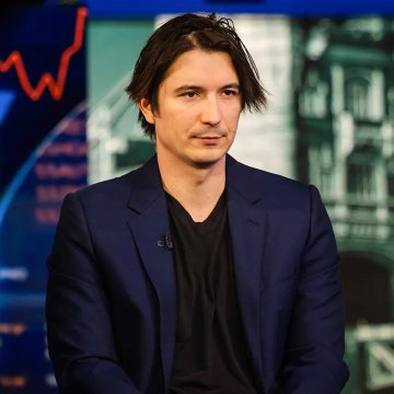 A man with dark hair wearing a navy blue blazer over a black shirt appears on what looks like a financial news broadcast set, with a downward red trend line visible in the background—suggesting market discussion. The image likely relates to news about Robinhood Chain's $400 million in asset deposits, a 300% weekly increase according to Token Terminal, possibly tied to broader crypto market movements.