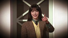 A young woman with bangs smiles and waves at the camera while wearing a brown blazer over a yellow sweater vest and white shirt.