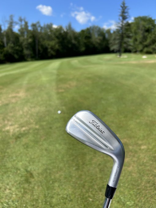 A close-up view shows a Titleist golf wedge on a grassy fairway with a golf ball visible in the distance.