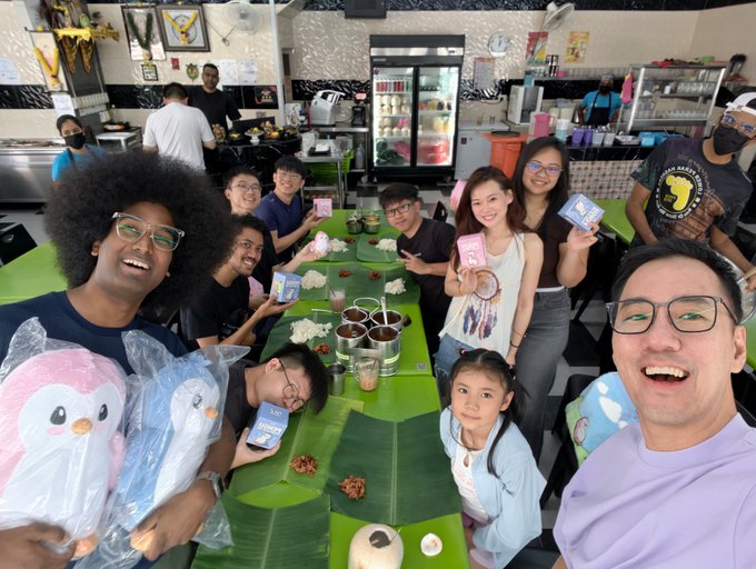 Group selfie taken at a restaurant table covered with banana leaves, showing a diverse group of young people smiling and holding packaged plush toys.