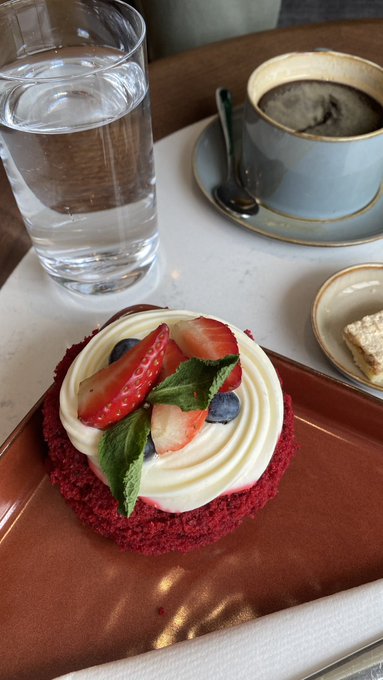 A photo of a red velvet cupcake topped with cream, fresh strawberries, blueberries, and mint leaves on a brown plate, accompanied by a glass of water and a cup of coffee on a tray.