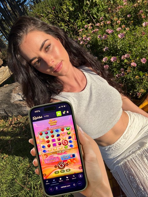 A woman with long dark hair poses outdoors against a backdrop of pink flowers and greenery while holding up a smartphone displaying the Rainbet.com mobile casino site, featuring the Sweet Bonanza slot game with a balance of $39.30 and a $39 spin bet amount visible on screen. The image visually promotes her post offering to spin Sweet Bonanza with her $39 balance.