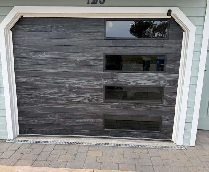A modern dark gray wood-grain garage door featuring horizontal paneled windows set into a light green siding house, house number '120' partially visible above.
