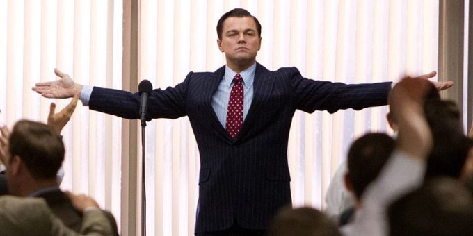 Leonardo DiCaprio stands triumphantly with arms outstretched in a navy pinstripe suit and red tie, evoking the iconic "Wolf of Wall Street" pose as he addresses an adoring crowd, while the post's text about conviction and diamond hands on $UFD appears as a humorous juxtaposition with the scene.