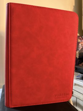 A red leather game master screen or binder stands upright on a dark surface, appearing to be a tabletop RPG accessory.