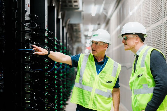 Two technicians in white hard hats and high-visibility vests branded with "EE" examine server racks in a data center, with one pointing at equipment while the other observes. The image visually reinforces the post's commentary on Neoclouds like $NBIS and $IREN, highlighting the tangible infrastructure—servers and skilled personnel—that underpins the scarcity of deliverable AI compute capacity.