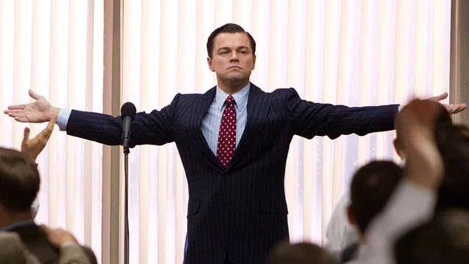 A man in a pinstripe suit and red polka-dot tie stands behind a microphone with his arms outstretched triumphantly before an applauding crowd. The image evokes the celebrity-like status of traders, capturing a moment of dramatic acclaim that mirrors how Wall Street figures often command rock-star attention and adoration.