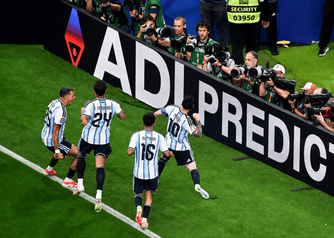 Lionel Messi (#10) and Argentina teammates Fernández (#24) and Almada (#16) run toward photographers after a goal, as Adidas-branded boards line the pitch—imagery used to visually echo the post's claim about ADI serving as a "settlement layer" for digital collaboration.