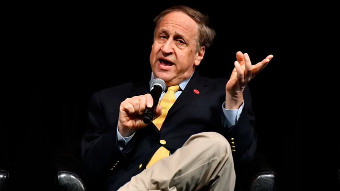 A man in a dark suit with a yellow tie speaking into a microphone while seated on stage against a black background, gesturing expressively with his left hand.