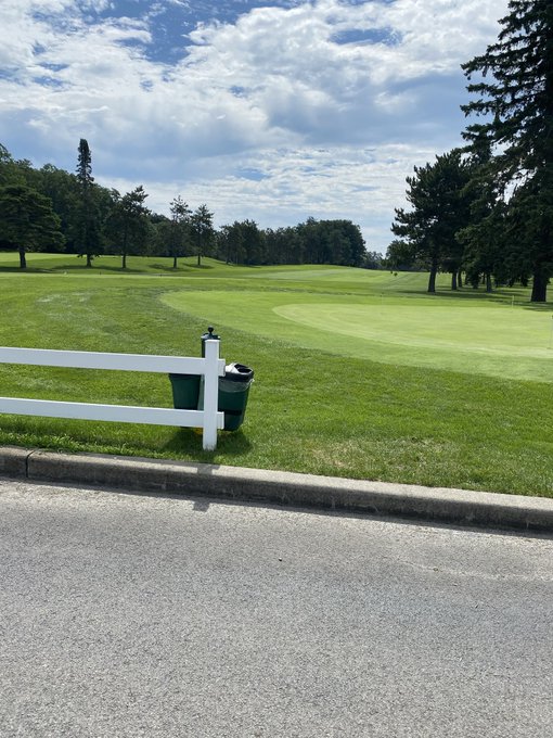 A scenic view of a golf course with green fairways and trees, seen from behind a white fence and green trash bins along a paved path under a partly cloudy sky.