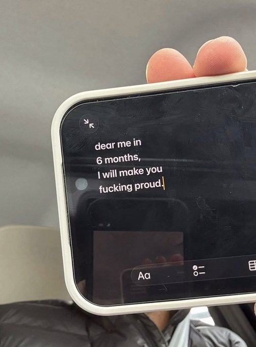 The image shows a person holding up their phone displaying a message that reads "Dear me, I will make you Fucking proud!". The person's fingers are visible as they hold the phone against the background of a gray wall. The text on the screen is in black and white, which contrasts with the rest of the image.