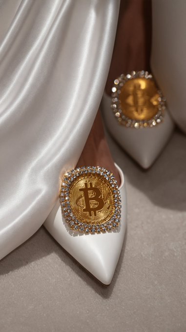 A close-up shows white pointed-toe high heels adorned with large gold Bitcoin coins surrounded by diamond-like crystals.