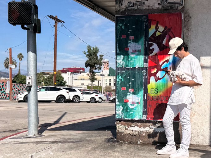 A photograph shows a young man dressed in all white clothing and a beige cap leaning against a graffiti-covered utility box at a street corner, focused on writing in a notebook with a street and white cars visible in the background.