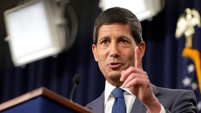 A man in a dark suit with a blue tie speaks at a podium, gesturing upward with his finger during what appears to be an official briefing, with a partial American flag visible in the background, tying the image to the post's focus on upcoming U.S. jobs data.