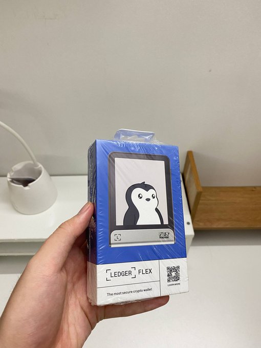 A hand holds a shrink-wrapped blue box for a 'LEDGER FLEX' crypto hardware wallet featuring a penguin illustration, with text reading 'The most secure crypto wallet'.