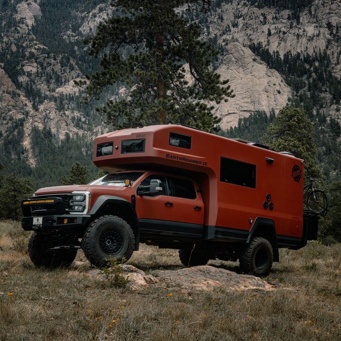 A rugged orange EarthRoamer LT expedition vehicle parked on rocky terrain with pine trees and a mountain cliff face in the background.