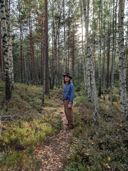 Portrait photograph of a person wearing a cowboy hat, blue button-up shirt, and brown pants standing on a forest path surrounded by tall pine and birch trees with sunlight filtering through.