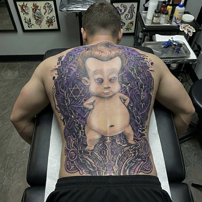 A man's back displays an elaborate full-back tattoo featuring a baby figure surrounded by purple patterns, chains, and astrological symbols in a tattoo parlor.